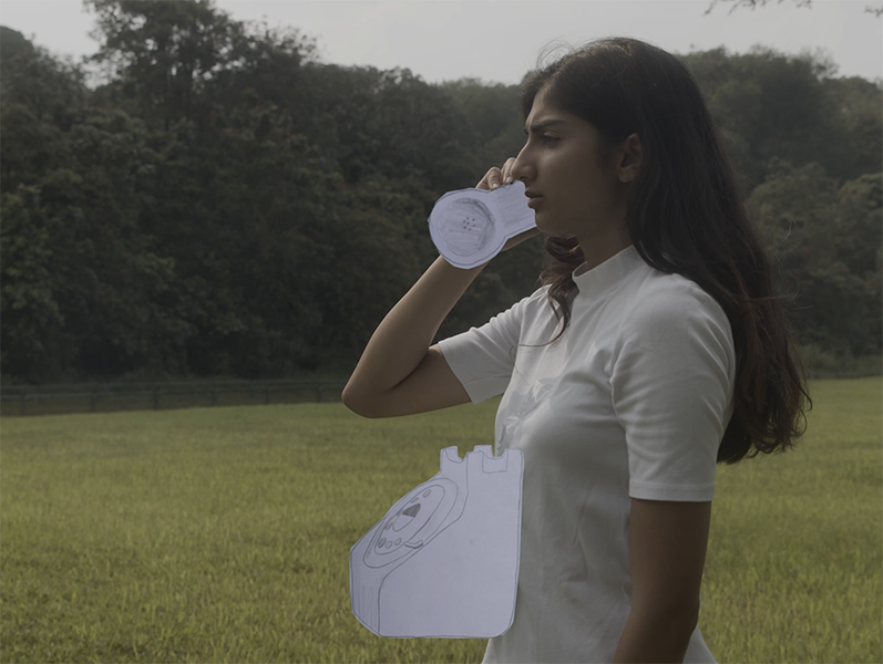 A woman with long hair in a white tshirt stands in a field, holding a paper cut outphone receiver. The paper rotary telephone body is attached to her stomach.