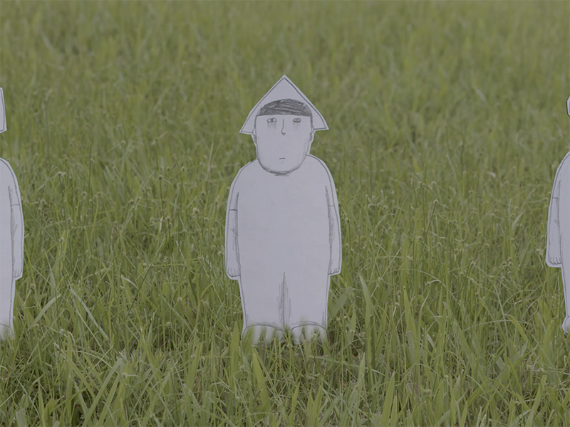 A paper cut out tiny man with a triangle shaped hat stands in a grass field staring into the camera.