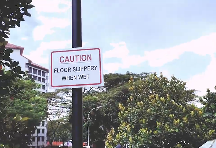 A photographic image of a caution: floor slippery when wet sign on a pole, with trees and a residential block in the background